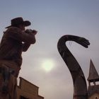 Ein Cowboy zielt mit einem Gewehr auf eine große, aufgerichtete Schlange vor einem Sonnenuntergang und einem Kirchturm.