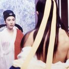 Zwei Personen in traditioneller asiatischer Kleidung; eine trägt einen roten Umhang und einen schwarzen Hut, die andere ein Band im Haar.