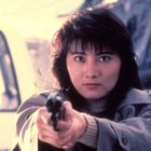 Eine Frau mit konzentriertem Blick zielt mit einer Pistole in Richtung Betrachter. Hintergrund leicht verschwommen.