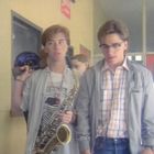 "Exit" steht auf einem Schild über einer Tür. Zwei junge Personen: Eine trägt ein Saxophon und ein Helm, die andere eine Brille.