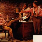 Drei Männer in Westernkleidung, in einer rustikalen, mit Ziegeln ausgekleideten Umgebung. Einer lehnt am Tisch, ein anderer sitzt entspannt.