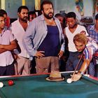 Ein Mann zielt konzentriert mit einem Queue auf einem Billardtisch; um ihn herum stehen mehrere Leute aufmerksam.