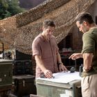 Zwei Männer betrachten eine Karte auf einem Tisch, umgeben von militärischen Kisten und einem Tarnnetz im Hintergrund.