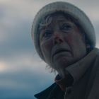 Ein älterer Mensch mit weißer Mütze und Wintermantel schaut besorgt in die Ferne vor bewölktem Himmel.