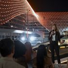"Vanderberg Motorsport", Rennwagen, Frau spricht mit Mikrofon, Publikum, Hintergrund beleuchtet, moderne Architektur bei Nacht.