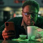 Ein Mann schaut auf sein Handy, trinkt mit einem Strohhalm aus einem Becher. Fast Food Verpackungen vor ihm.