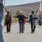 Drei Männer in Polizeiuniformen laufen auf einer leeren Straße, einer trägt einen Cowboyhut. Im Hintergrund eine Berglandschaft.