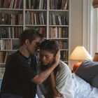 Ein Paar sitzt auf einem Bett, umarmend. Hintergrund: Bücherregal, Kamin, Lampe und Fenster mit Bäumen draußen.