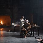 Zwei Personen in einem düsteren Raum; einer sitzt, der andere macht ihm Gesichtspflege, vor einem prasselnden Kaminfeuer.