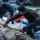 Zwei Menschen auf einem schneebedeckten Ufer, eine Person wird gestrichen, umgeben von zwei helfenden Personen und einem roten Kanu.