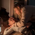 Zwei Personen auf dem Boden; eine liegt, die andere kniet besorgt daneben. Eine Lampe beleuchtet sanft die Szene.