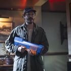 Ein Mann hält eine Wasserpistole mit der Aufschrift "Psycho Hydro" in einem gemütlichen Raum mit Gemälde und Pflanzen.