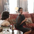 Zwei Frauen sitzen auf einem roten Sofa mit Mustern, trinken Tee. Im Hintergrund steht eine Ritterrüstung in einem eleganten Raum.