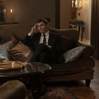 Ein Mann in Anzug sitzt nachdenklich auf einem Sofa in einem luxuriös eingerichteten Raum mit Kerzen und Kissen.