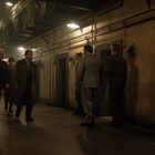 Mehrere Männer in Militäruniformen und Mänteln gehen durch einen schummrigen, industriellen Korridor mit schweren Türen.