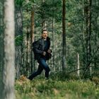 Ein Mann in Outdoor-Kleidung joggt durch einen dichten Wald, umgeben von hohen Bäumen und grünem Laub.
