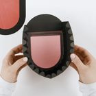 Keine Texte vorhanden. Hände halten ein schwarz-rosa geometrisches Objekt mit gezackten Rändern auf weißem Hintergrund.