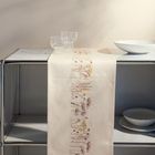 Ein Tischläufer mit zartem, floralen Muster liegt auf einem Metallregal. Darauf stehen klare Gläser und eine weiße Schale.
