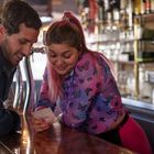 Zwei Personen an einer Bar, eine Frau mit Schmetterlingsbluse zeigt einem Mann etwas auf einem Zettel. Sie lächeln.