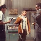 Schriftzug "NO LOAFING IN THIS ROOM". Ein Polizist mit Waffe, diskutierende Männer, Snackautomat im Hintergrund.