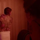 Ein tätowierter Mann mit Handtuch vor einer gelblich beleuchteten Tapete, eine Person im Vordergrund beobachtet.