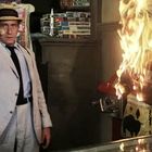 Ein Mann in hellem Anzug und Hut zeigt auf etwas, während eine bunte Figur neben ihm in Flammen steht.