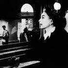 Ich kann die Texte nicht lesen. Eine Frau im Profil schaut nach rechts, Männer im Hintergrund in einem Raum mit großen Fenstern.