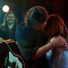 Ein Paar umarmt sich auf einer Bühne, während ein Musiker im Hintergrund lächelt. Eine Gitarre ist sichtbar.
