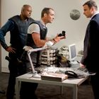 Ein Mann in einer Schutzweste hält ein Objekt, zwei andere Männer schauen zu. Im Raum steht ein Tisch mit Computer und Lampe.