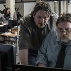 Zwei Männer in einem Büro schauen konzentriert auf einen Computerbildschirm. Im Hintergrund arbeiten weitere Personen.