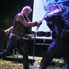 Es gibt keine lesbaren Texte. Zwei Männer kämpfen mit Gitarren auf einer Bühne voller Konfetti. Eine Schlagzeugausrüstung ist im Hintergrund.