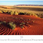 "Düne aus rotem Sand", und es zeigt eine Wüstenlandschaft mit roten Dünen und Gräsern. Im Hintergrund sind Berge.