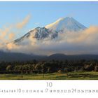 „Mount Egmont“ steht unten links. Es zeigt einen schneebedeckten Berg mit Wolken davor und bewaldetem Vordergrund.