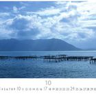 "Morgenstimmung am Strand." Kalenderblatt mit Meer, Holzstegen und Bergen im Hintergrund unter einer bewölkten Himmel.
