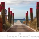 "Weg zum Meer", darunter ein hölzerner Steg von Dünen flankiert, führt zum Meer, eingerahmt von rot bemalten Pfählen.