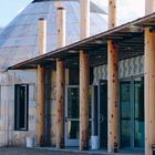 Großes rundes Gebäude mit Holzkonstruktionen und modernen Türen. Der Eingang ist durch Säulen gestaltet.