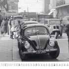 Viele Schilder mit Namen von Geschäften; Straßenszene der 1950er Jahre mit geparktem schwarzen Käfer im Vordergrund.