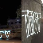 Links Graffiti „EROS“, rechts ein Zug in Bewegung in einer U-Bahnstation. Gebäudefassade im Hintergrund.