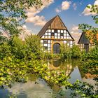 Ein Fachwerkhaus mit dunklem Holzdach spiegelt sich in einem Teich, umgeben von üppigen grünen Bäumen und blauem Himmel.