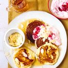 Pfannkuchen mit Mandeln und Sirup auf weißem Teller, daneben Schüssel mit Joghurt und Beeren, goldener Hintergrund.