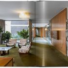 Ein heller Raum mit grünen Teppich, zwei Sesseln und einem weißen Tisch, umgeben von Pflanzen und Holzverkleidung.