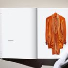 Text: "Club Poetry Gold Lame Suit". Orangefarbener Anzug in Buch, gehalten von einer Hand mit weißem Handschuh.