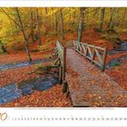 Kalender zeigt Oktober 2023. Holzbogenbrücke über einen Bach in einem herbstlichen Wald mit orangefarbenem Laub.