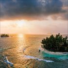 Keine Texte erkennbar. 

Kleine Insel mit Palmen, umgeben von türkisfarbenem Wasser; Sonnenuntergang am Horizont.üstere Wolken.