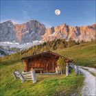 Eine Holzhütte steht auf einer Wiese, umgeben von einem Zaun. Dahinter erheben sich beeindruckende Berge bei Vollmond.