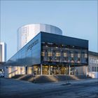 Text: "PERGAMON" und "PERGAMON PANORAMA". 

Beschreibung: Modernes Gebäude mit Glasfassade, Lichtern und Treppen.