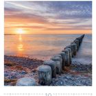 Text: "01-31 Oktober". Steinküste mit Holzpfählen in der Brandung, Sonnenuntergang am Horizont, sanfte Wolken am Himmel.