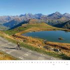 "Oktober" steht groß unten. Radfahrer auf Bergstraße, Berge und See im Hintergrund. Logo unten rechts.