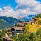 Es gibt keine sichtbaren Texte. Eine malerische Berglandschaft mit Holzhäusern, grünen Hügeln und einem Kirchturm im Zentrum.