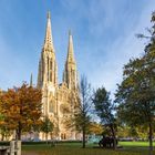 Spitze Türme einer gotischen Kathedrale, umgeben von Bäumen mit buntem Herbstlaub unter einem klaren blauen Himmel.
